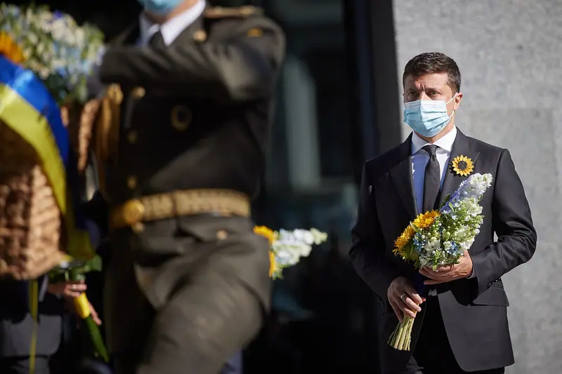 A man in a dark suit and a light blue face mask holds a bouquet of white and blue flowers with sunflowers, while a uniformed honour guard in the blurred foreground carries a wreath with a blue and yellow ribbon