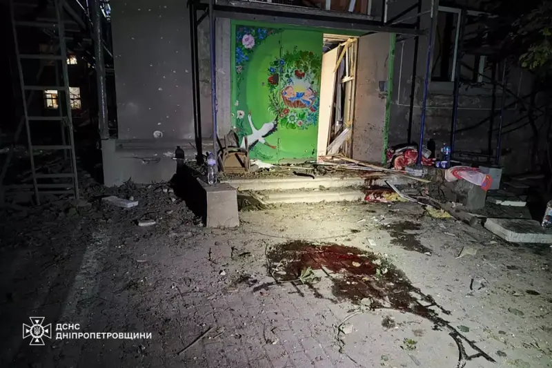 Night view of a hospital entrance with a painted door blown open, rubble and debris on the steps and a pool of blood on the paving