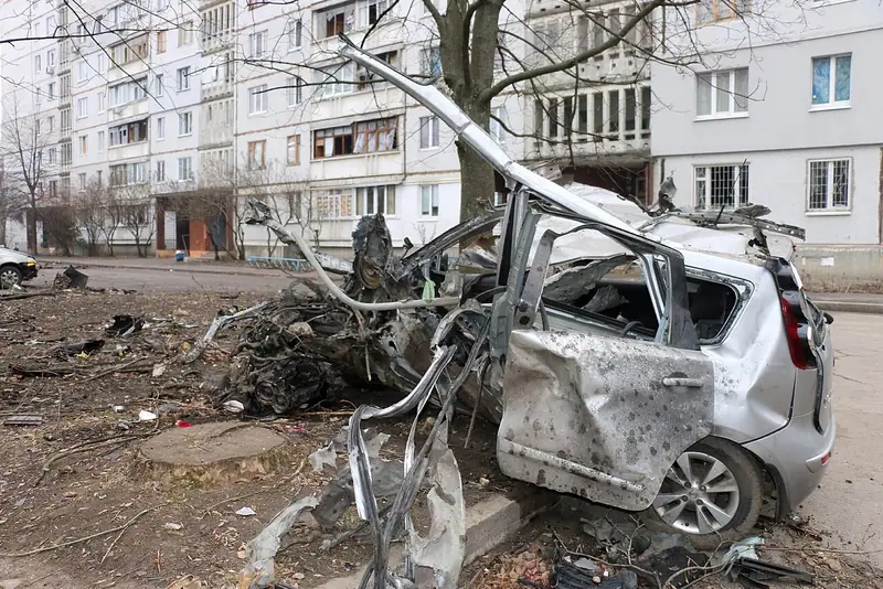A wrecked, shrapnel-pierced car with its roof torn open in front of damaged apartment blocks in Northern Saltivka