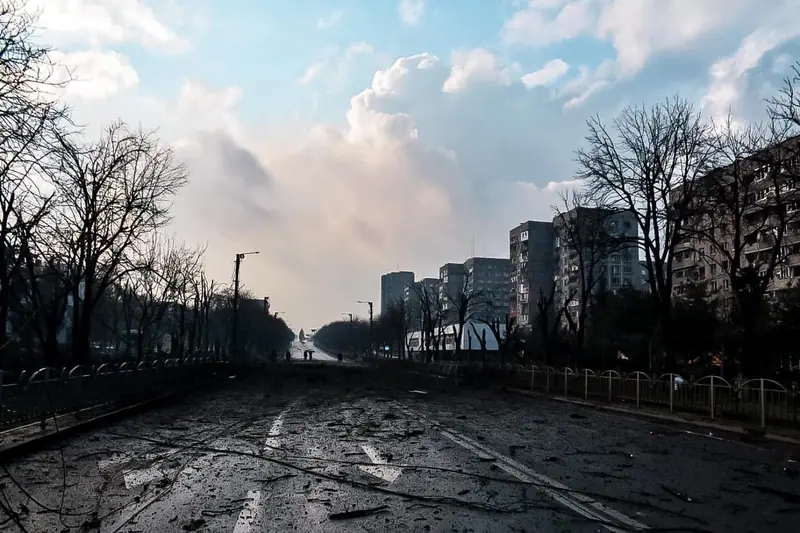 A wide avenue in Mariupol strewn with debris and fallen cables between apartment blocks, with a large cloud of smoke rising in the distance