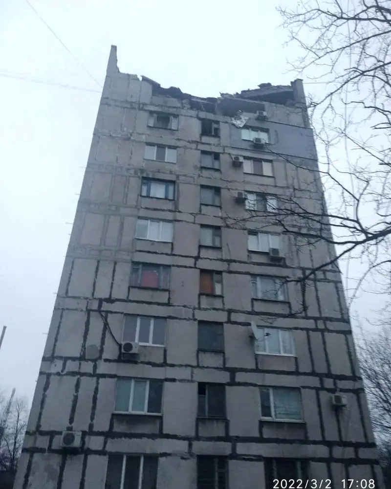 The upper floors of a nine-storey concrete apartment block with part of the roof edge torn away and many windows missing, seen from below, with a date stamp of 2 March 2022