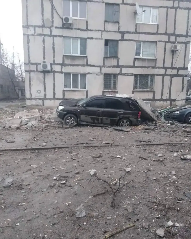 A dark SUV and a second car beside an apartment block, with a slab of concrete fallen onto the rear of the SUV and debris covering the ground