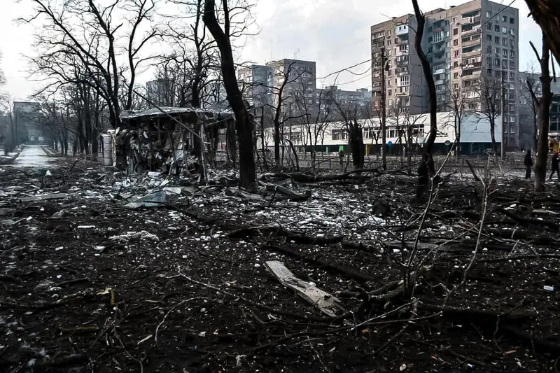 A park or street area in Mariupol covered in broken branches and debris, with a wrecked kiosk and apartment blocks in the background