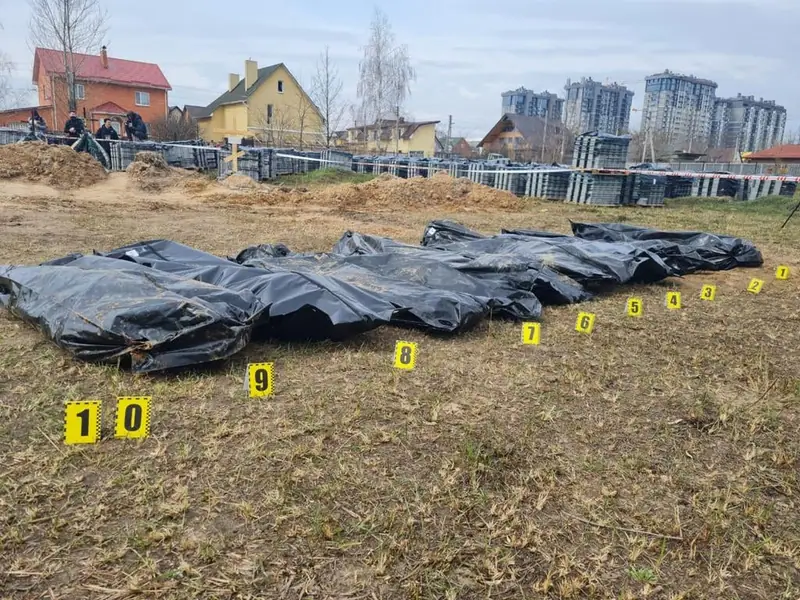 Ten black body bags laid in a row on dry grass, each marked with a numbered yellow evidence tag, with fenced grave sites and apartment blocks in the background