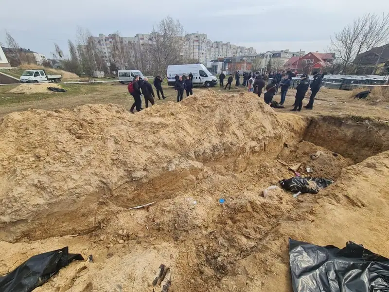 A large open pit and mounds of sand with black plastic sheeting in it, and a group of police officers and investigators standing behind it next to white vans