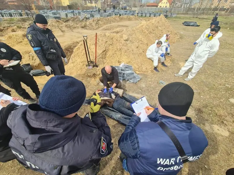 Investigators in white protective suits and police officers around a body lying on a black sheet beside a mound of excavated earth, one wearing a jacket marked "War Crimes Prosecutor"