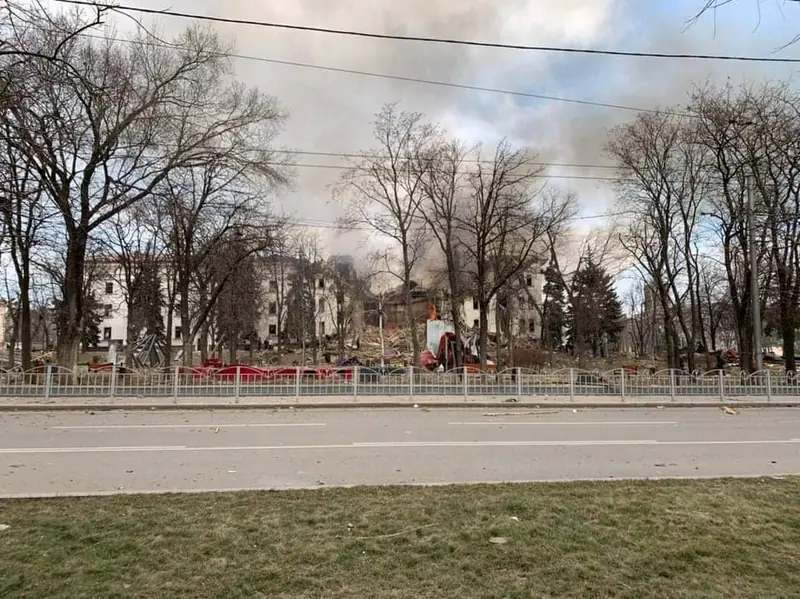 Smoke rises behind bare trees over the wrecked Mariupol Drama Theatre, with debris and damaged cars in front of it