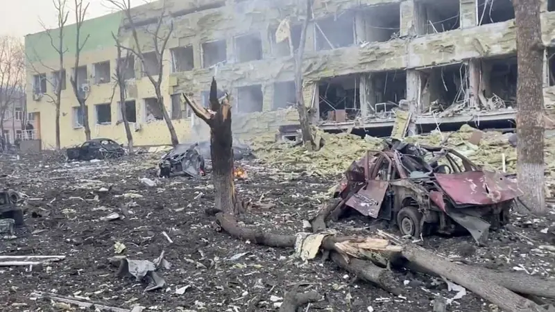 A heavily damaged multi-storey hospital building with blown-out walls and windows, a burnt-out car and broken trees in the debris-covered courtyard