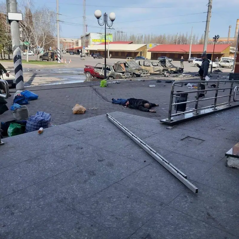 The station forecourt with luggage bags and a plastic bag on the ground, one person lying on the paving, several burnt-out cars on the road behind, and a man standing by the railing