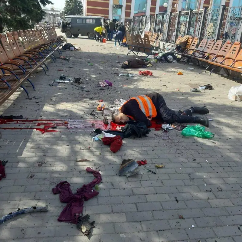 Station platform area with wooden benches, scattered belongings, clothing and a large pool of blood, with a person lying on the paving and rescuers standing near a van in the background