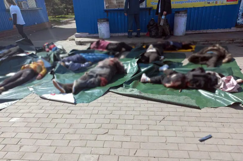 Rescuers standing beside rows of bodies laid on green tarpaulins on the paving outside a blue kiosk, with the bodies blurred in the published photo