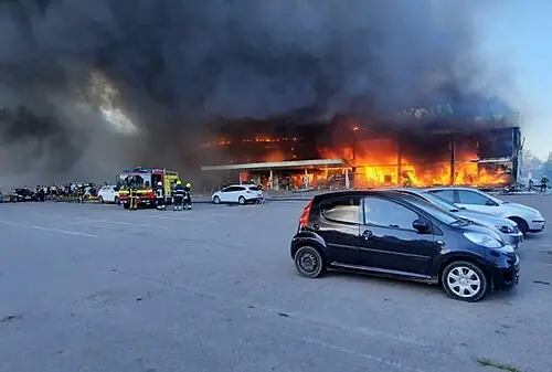Flames and thick black smoke pour from the Amstor shopping centre while firefighters and a fire engine stand by. Parked cars in the foreground