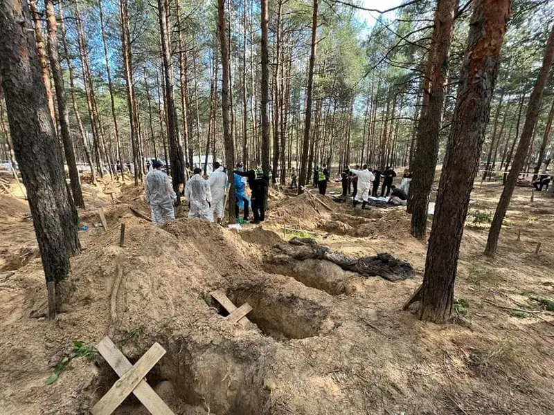 Investigators in white protective suits standing among open and filled graves marked with wooden crosses in a pine forest