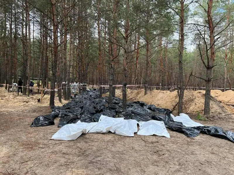 Many black and white body bags laid out on the sandy ground in a pine forest next to a long excavated trench, with police tape and a few people in the background