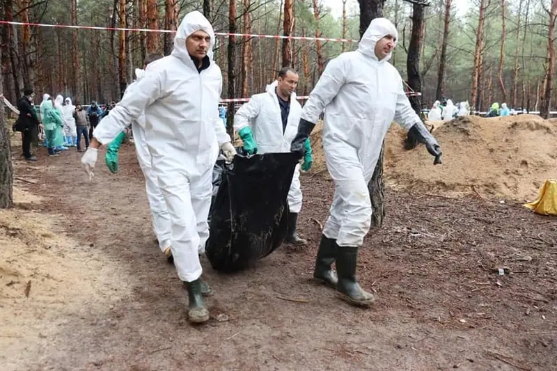Three people in white protective suits and rubber boots carrying a black body bag along a forest path, with others working behind police tape