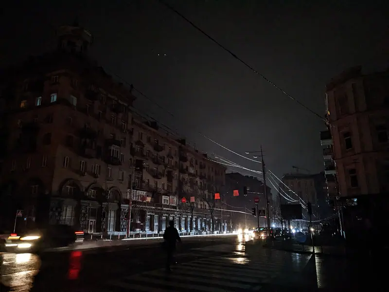 A wide Kyiv street at night with dark apartment buildings and no streetlights, lit only by car headlights