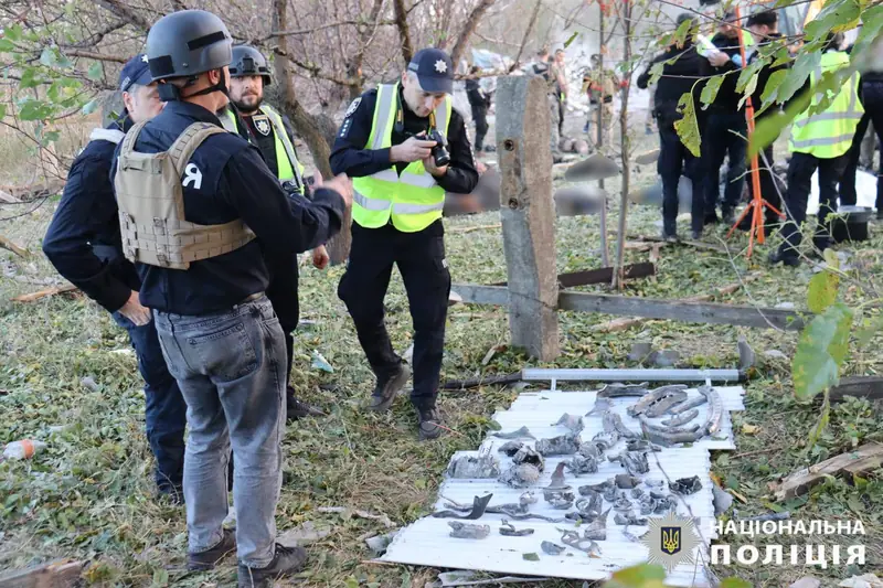 Police investigators in vests and helmets, one photographing, standing over a white metal sheet on which fragments of a missile are laid out in rows, with other officers working in the background