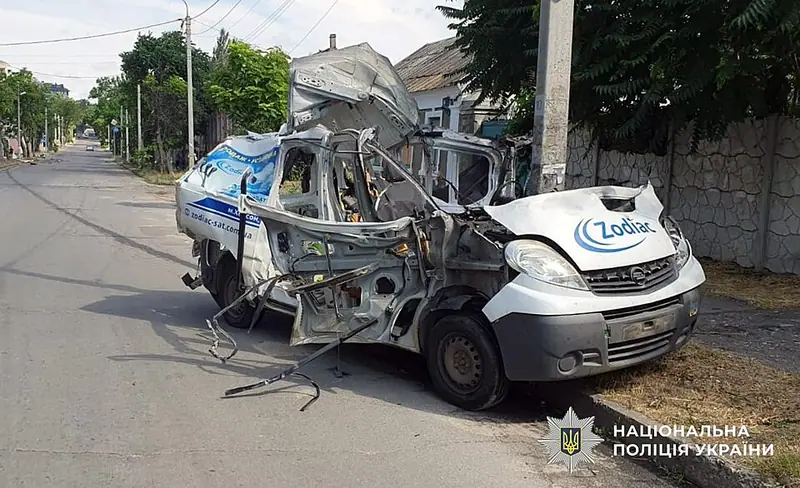 A white van with its side and roof torn open and debris on the road beside it, parked by a kerb on a tree-lined street