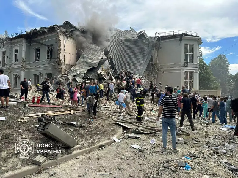A crowd of rescuers, medical staff and volunteers climbing over rubble in front of a hospital building whose roof has collapsed, with smoke rising