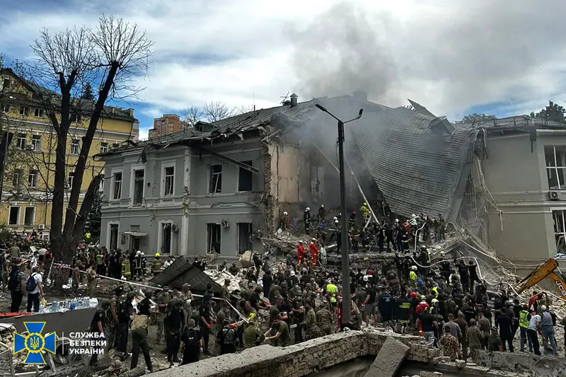 A very large crowd of soldiers, police, firefighters and civilians gathered at the rubble of a collapsed hospital wing, with smoke rising behind a grey two-storey building