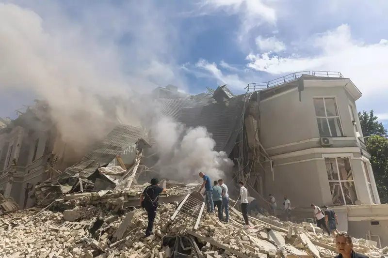 A small group of people in civilian clothes and a police officer standing on a pile of bricks and broken masonry in front of a collapsed roof, with thick smoke behind them