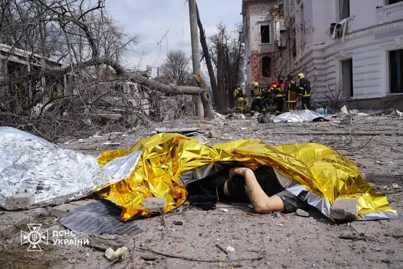 A body covered with a gold and silver emergency blanket on a rubble-strewn street, with firefighters and a damaged building in the background