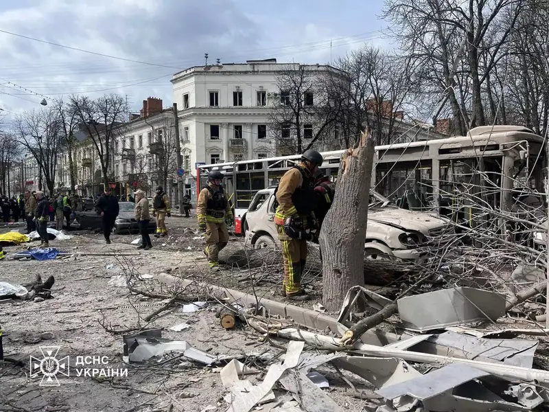 Firefighters walking among broken tree trunks and debris on a city street, with a damaged trolleybus, a wrecked car and blown-out windows of buildings behind them
