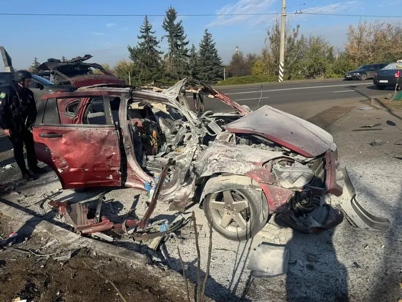 The burned-out and torn open remains of a red car on a road, with a police officer in a helmet standing beside it