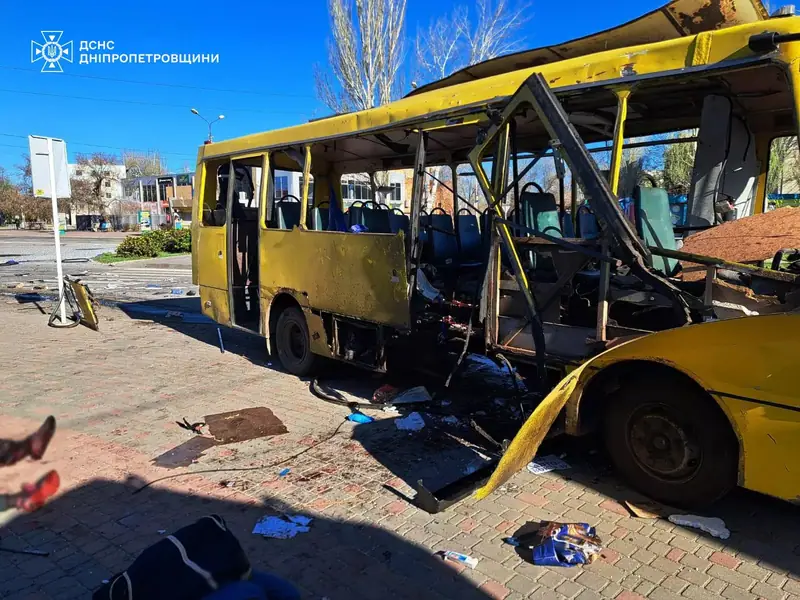 A yellow minibus at a stop with its roof torn off, its side and front destroyed, blood on the paving, and a person's feet visible at the left edge