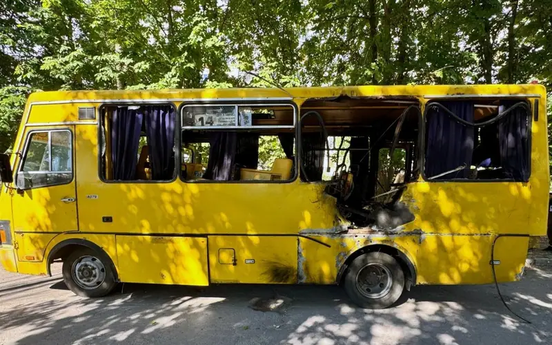 A yellow minibus with blown-out windows and a burnt, torn side panel parked under trees