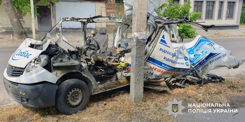 A white van with its cab and rear section torn apart and its roof peeled open, stopped against a concrete pole beside a road