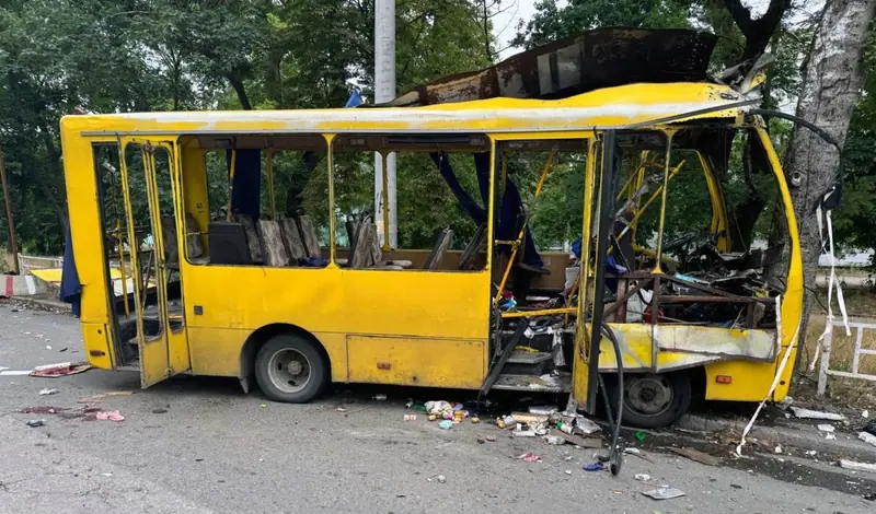 A yellow minibus with its roof torn open, front destroyed and doors blown out, with blood on the road beside it