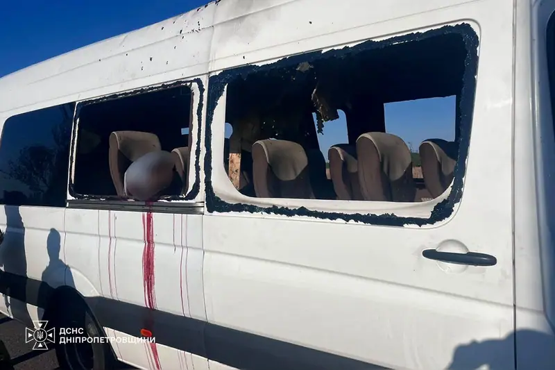 A white minibus with both side windows blown out and pitted with shrapnel marks, blood running down its side below a blurred area at one window