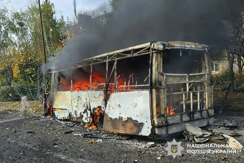 A burned-out bus frame with flames still burning inside and black smoke rising, on a street lined with trees in Kramatorsk.