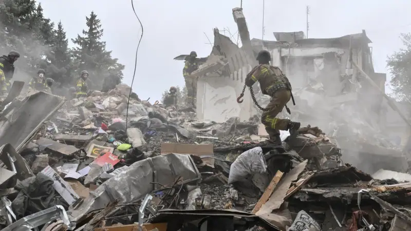 Rescuers in helmets climb over a large pile of rubble and household belongings in front of the collapsed apartment building, one carrying a chain.